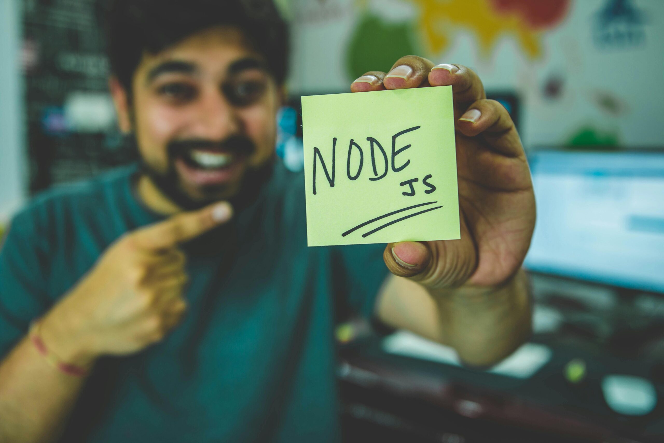 A cheerful software developer shows a Node.js sticky note in a tech workspace.